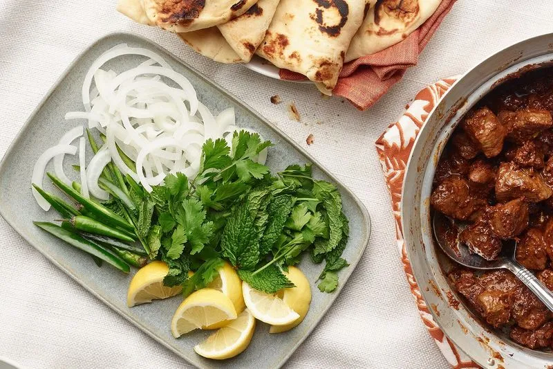 Fresh herbs, sliced onion and lemon beside a pan of seasoned meat and warm flatbread